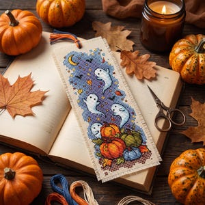 Peut inclure: Un marque-page rectangulaire en tissu orné d'un motif au point de croix représentant trois fantômes blancs, des citrouilles orange et vertes, un croissant de lune jaune et des petites chauves-souris sur fond violet, avec un pompon bleu et orange.