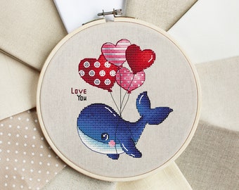 Könnte beinhalten: Stickrahmen mit einer Kreuzstich-Stickerei, die eine blaue Wal, die herzförmige Ballons hält, zeigt. Die Ballons sind rot, rosa und weiß mit Punkten und Streifen. Der Text "Love You" ist unter dem Wal gestickt.