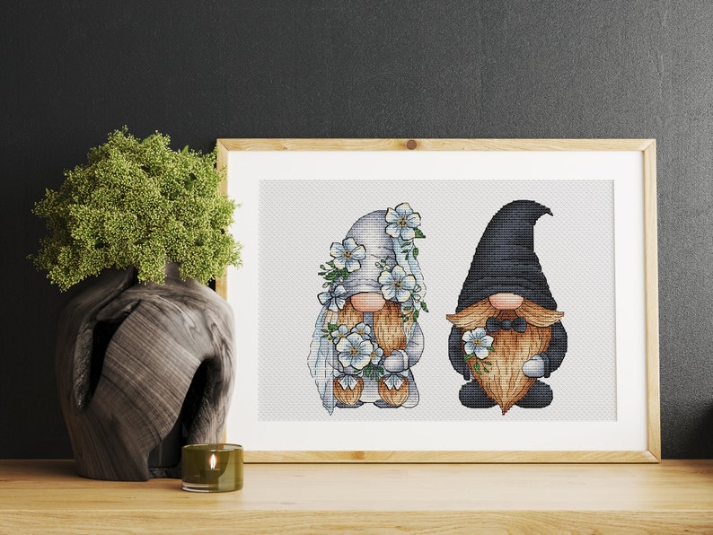 May include: A cross-stitch pattern featuring a bride and groom gnome couple. The bride gnome wears a white dress and veil with blue flowers. The groom gnome wears a black suit with a bow tie and holds a bouquet of blue flowers.