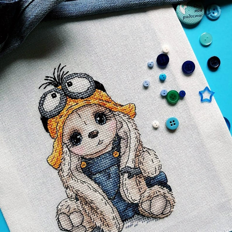 K&ouml;nnte beinhalten: Ein Kreuzstichmuster eines niedlichen Kaninchens, das einen gelben Fliegerhut mit Brille und eine blaue Jeansjacke tr&auml;gt. Das Kaninchen sitzt mit gekreuzten Pfoten und hat einen s&uuml;&szlig;en Gesichtsausdruck.