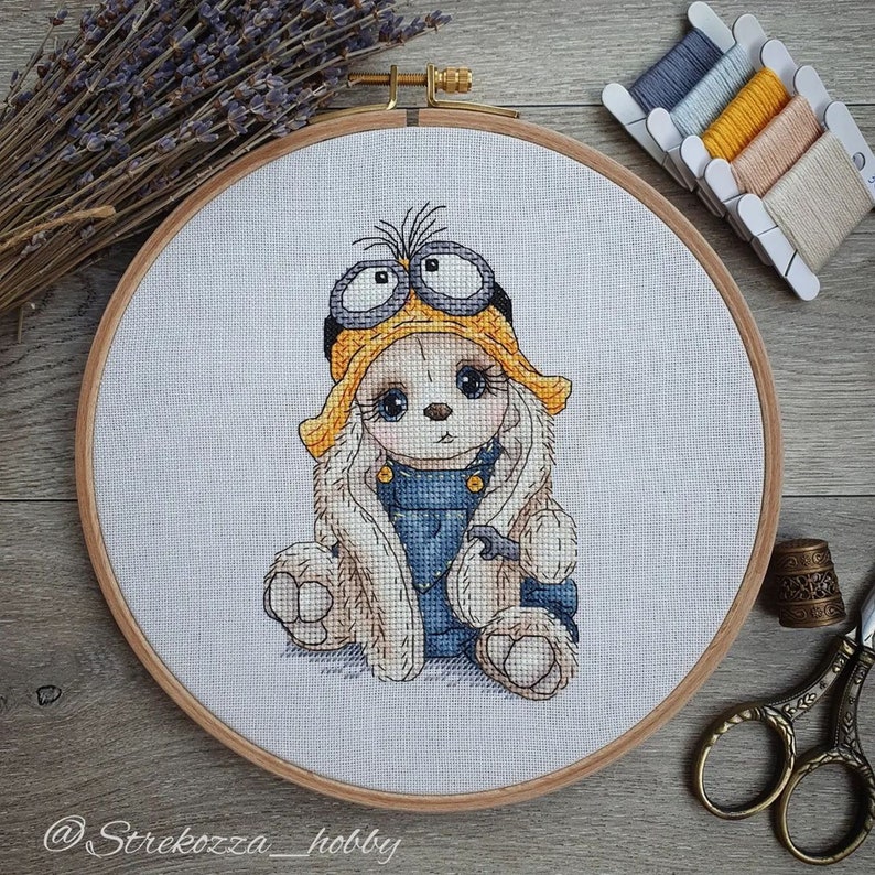 K&ouml;nnte beinhalten: Kreuzstich-Stickerei eines wei&szlig;en Kaninchens, das einen gelben Hut mit Schutzbrille und einen blauen Jeans-Overall tr&auml;gt. Die Stickerei ist in einem Holzreifen gerahmt.
