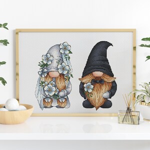 May include: Cross-stitch pattern of a bride and groom gnome couple. The bride gnome is wearing a white dress with blue flowers and a veil. The groom gnome is wearing a black suit with a bow tie and a black hat.