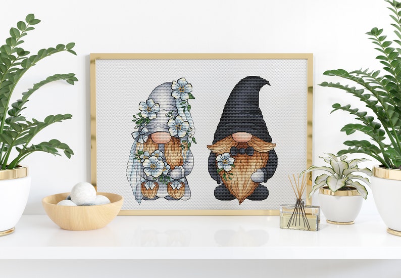 May include: Cross-stitch pattern of a bride and groom gnome couple. The bride gnome is wearing a white dress with blue flowers and a veil. The groom gnome is wearing a black suit with a bow tie and a black hat.