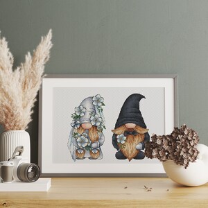 May include: A cross-stitch embroidery of two gnomes, one wearing a white hat and veil with blue flowers, the other wearing a black hat and a bow tie. Both gnomes are holding bouquets of blue flowers.