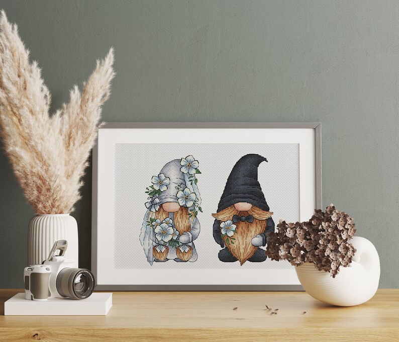 May include: A cross-stitch embroidery of two gnomes, one wearing a white hat and veil with blue flowers, the other wearing a black hat and a bow tie. Both gnomes are holding bouquets of blue flowers.