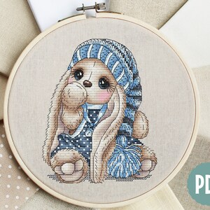 Puede incluir: Patr&oacute;n de punto de cruz de un lindo conejito con un gorro azul y blanco a rayas y un su&eacute;ter azul y blanco con lunares. El conejito sostiene un pomp&oacute;n azul.