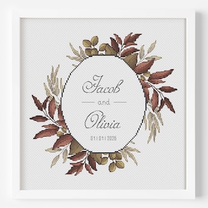 May include: A framed cross-stitch artwork featuring a circular design with the names "Jacob" and "Olivia" and the date 01/01/2026. The names are centered within a geometric shape, surrounded by a wreath of brown and gold leaves.