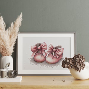 May include: A framed cross-stitch artwork featuring a pair of pink baby shoes with large bows. The shoes are detailed with cross-stitch patterns and sit on a white lace-like design. The artwork is displayed on a wooden surface.