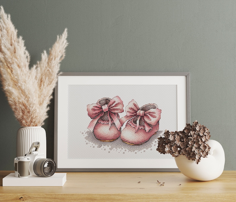 May include: A framed cross-stitch artwork featuring a pair of pink baby shoes with large bows. The shoes are detailed with cross-stitch patterns and sit on a white lace-like design. The artwork is displayed on a wooden surface.