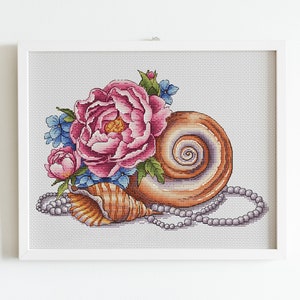 Könnte beinhalten: Ein Kreuzstich-Stickmuster mit einer Muschel mit einer rosa Pfingstrose und blauen Blumen, umgeben von einer Perlenkette.