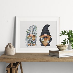 May include: A framed cross-stitch design featuring two gnomes, one in a white dress with blue flowers and the other in a black hat and beard, holding a bouquet of blue flowers.