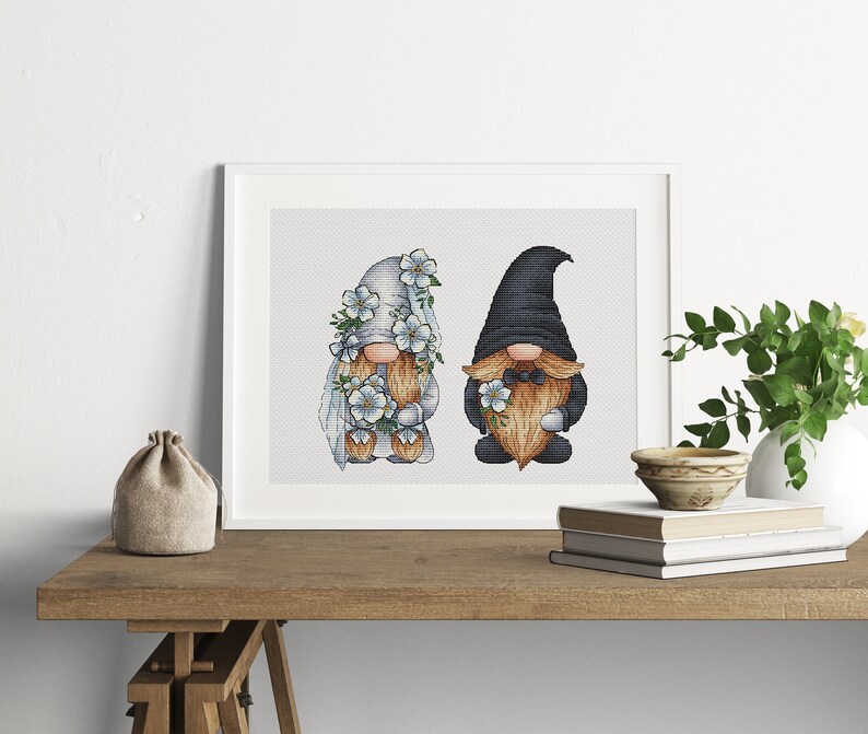 May include: A framed cross-stitch design featuring two gnomes, one in a white dress with blue flowers and the other in a black hat and beard, holding a bouquet of blue flowers.
