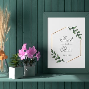 May include: A framed wedding announcement with a gold hexagonal frame and green foliage. The text reads "Jacob and Olivia 01.11.2023". The frame is on a shelf with a vase of orange tulips, a vase of dried wheat, and two potted plants.