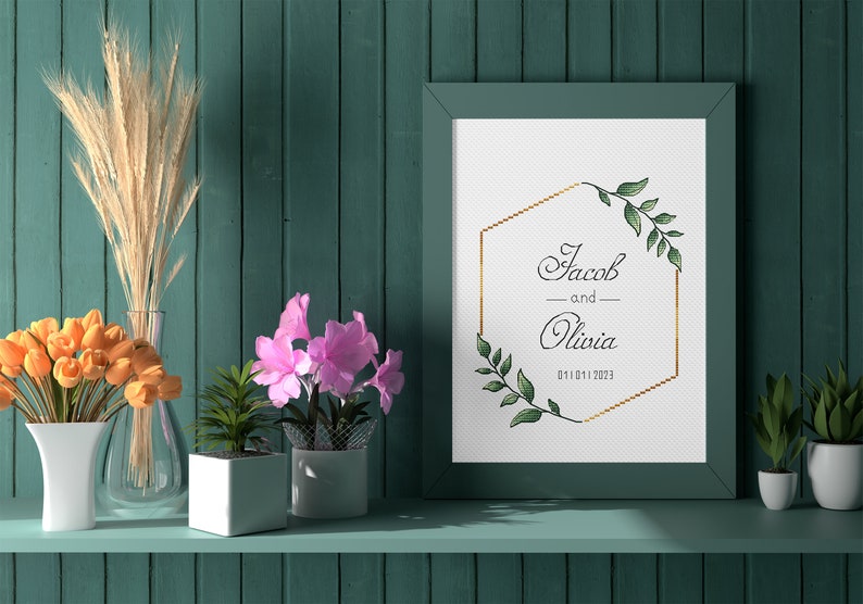 May include: A framed wedding announcement with a gold hexagonal frame and green foliage. The text reads "Jacob and Olivia 01.11.2023". The frame is on a shelf with a vase of orange tulips, a vase of dried wheat, and two potted plants.
