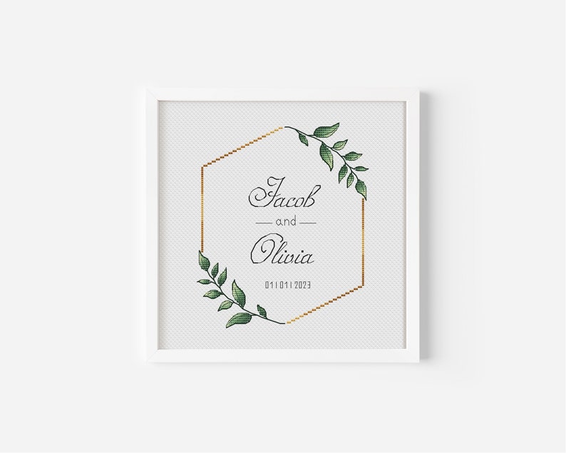 May include: A white framed print with a gold hexagonal frame and green leafy branches on each side. The text inside the frame reads "Jacob and Olivia 01.01.2023".