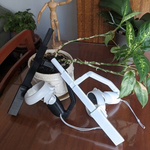 May include: Black and white VR controller accessories. The black accessory is paired with a black controller, and the white accessory is paired with a white controller. The controllers are resting on a brown table, with a wooden figurine and potted plants in the background.