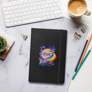May include: A black journal with a sticker featuring a colourful design and the text "ENERGY IS THE ONLY TRUE CURRENCY. SPEND YOURS WISELY." The journal is on a white desk with a keyboard, coffee, and pencils.