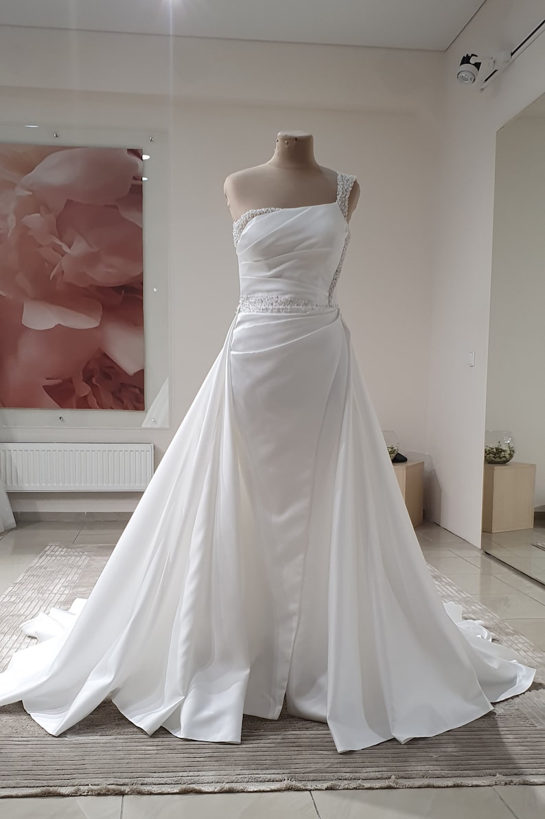 Puede incluir: Un vestido de novia de sat&eacute;n blanco con un dise&ntilde;o de un solo hombro y un corpi&ntilde;o con cuentas. El vestido tiene una falda fluida con cola.