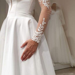 May include: A white strapless wedding dress with a full skirt and a lace overlay on one sleeve.