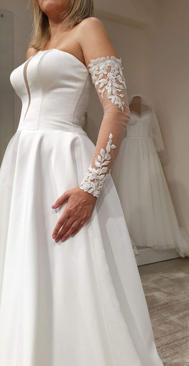 May include: A white strapless wedding dress with a full skirt and a lace overlay on one sleeve.