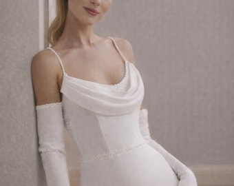 May include: A white wedding dress with a cowl neckline and spaghetti straps. The dress features a fitted bodice and a flowing skirt. Long, white gloves and a black bow complete the elegant bridal look.