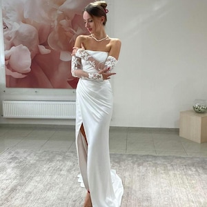 May include: A white off-the-shoulder wedding dress with a long, flowing skirt and a thigh-high slit. The dress features delicate lace detailing on the sleeves and a simple, elegant design.