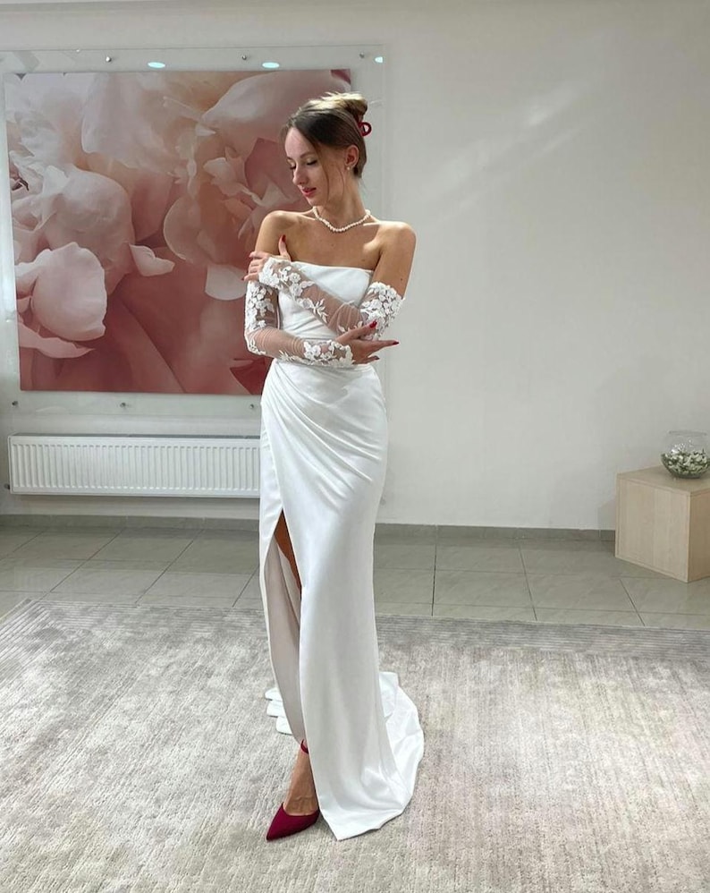May include: A white off-the-shoulder wedding dress with a long, flowing skirt and a thigh-high slit. The dress features delicate lace detailing on the sleeves and a simple, elegant design.