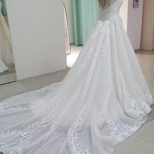 Puede incluir: Un vestido de novia blanco con un corpiño de encaje y una falda de tul. El vestido tiene una cola larga y está decorado con delicados bordados florales.