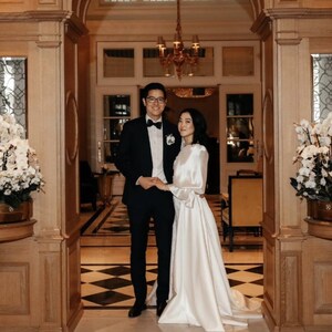 Puede incluir: Una pareja posa para una foto de boda en un gran pasillo ornamentado con un suelo de ajedrez. El novio lleva un esmoquin negro con pajarita, y la novia lleva un vestido blanco con una larga cola.