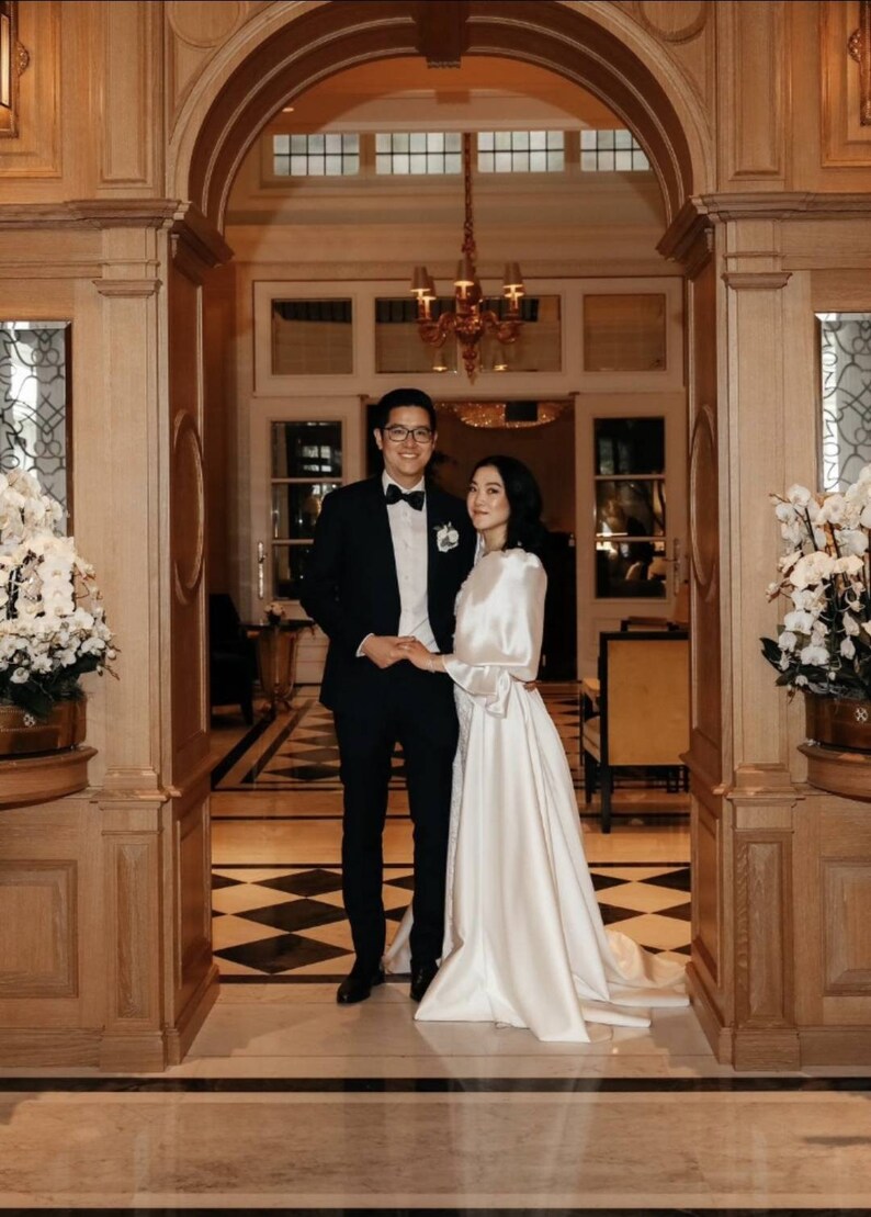 Puede incluir: Una pareja posa para una foto de boda en un gran pasillo ornamentado con un suelo de ajedrez. El novio lleva un esmoquin negro con pajarita, y la novia lleva un vestido blanco con una larga cola.