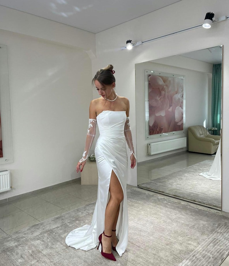 May include: A white strapless wedding dress with a thigh-high slit and lace detailing on the sleeves. The dress is fitted and has a flowing skirt. The model is wearing red heels.