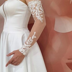 May include: White bridal gown with a strapless bodice and a full skirt. The gown features a sheer lace sleeve with floral embroidery.