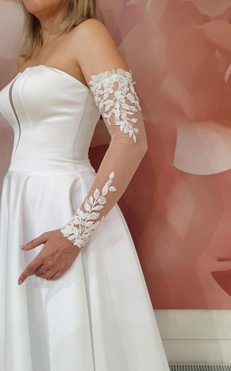 May include: White bridal gown with a strapless bodice and a full skirt. The gown features a sheer lace sleeve with floral embroidery.