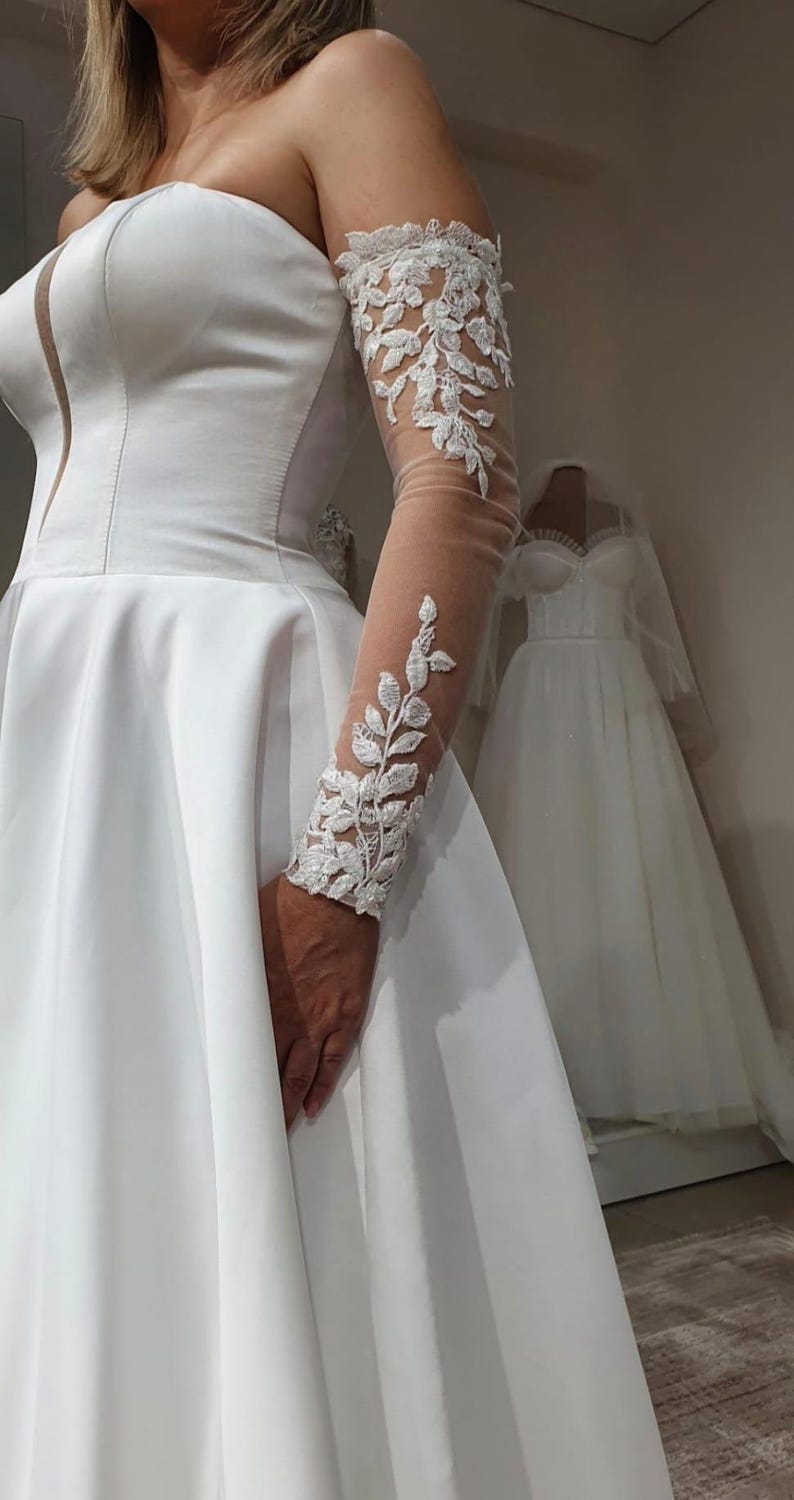 May include: A white strapless wedding dress with a full skirt and a lace overlay on the sleeves. The dress is made of a satin-like fabric and has a fitted bodice.