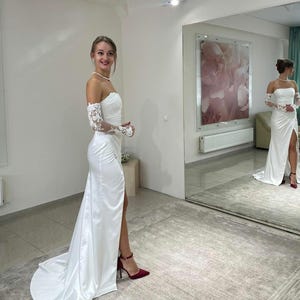 May include: A white satin wedding dress with a lace overlay on the sleeves and a thigh-high slit. The dress has a strapless bodice and a flowing skirt. The model is wearing red heels.