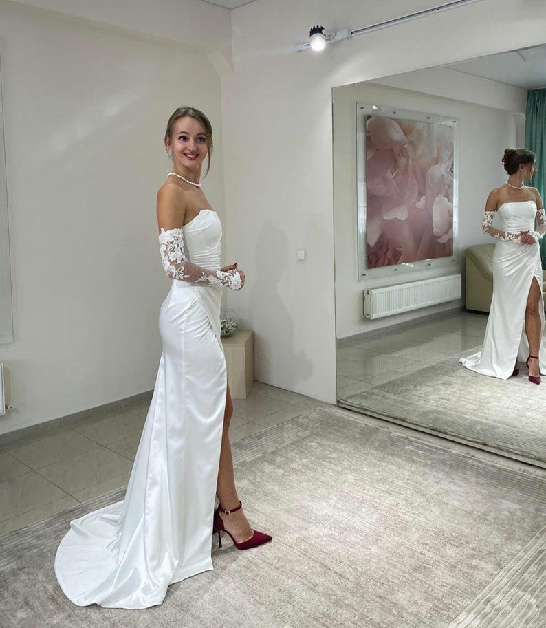 May include: A white satin wedding dress with a lace overlay on the sleeves and a thigh-high slit. The dress has a strapless bodice and a flowing skirt. The model is wearing red heels.