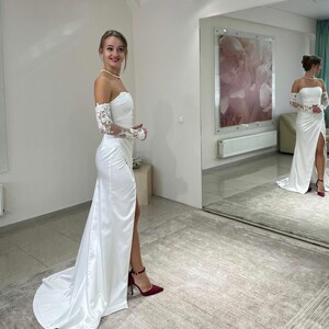 May include: A white satin wedding dress with a sweetheart neckline and a thigh-high slit. The dress has long, lace sleeves and a flowing train. The model is wearing red heels.