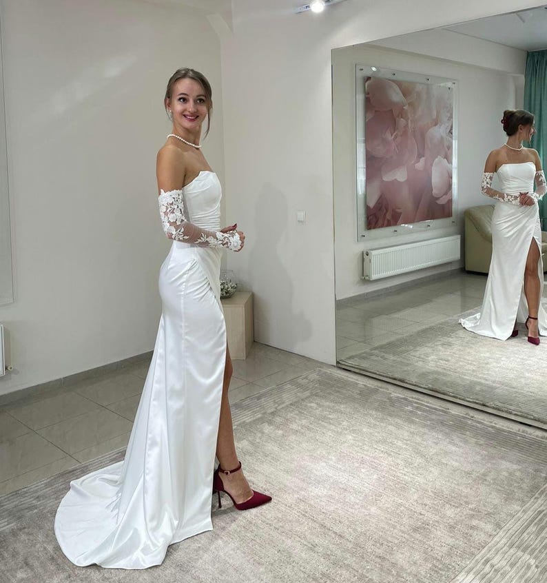 May include: A white satin wedding dress with a sweetheart neckline and a thigh-high slit. The dress has long, lace sleeves and a flowing train. The model is wearing red heels.
