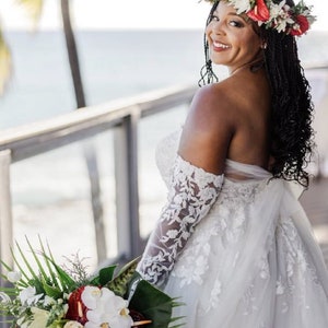 May include: A bride wearing a white off-the-shoulder lace wedding dress with a floral crown and holding a bouquet of tropical flowers.
