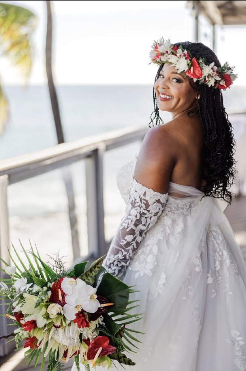 May include: A bride wearing a white off-the-shoulder lace wedding dress with a floral crown and holding a bouquet of tropical flowers.