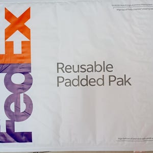 May include: White padded envelope with the FedEx logo in purple and orange. The envelope is labeled "Reusable Padded Pak".
