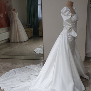 Puede incluir: Un vestido de novia de sat&eacute;n blanco con un corpi&ntilde;o ajustado y una falda fluida. El vestido tiene un escote de coraz&oacute;n y mangas largas con hombros abullonados. La falda tiene una cola y una sobrefalda desmontable.