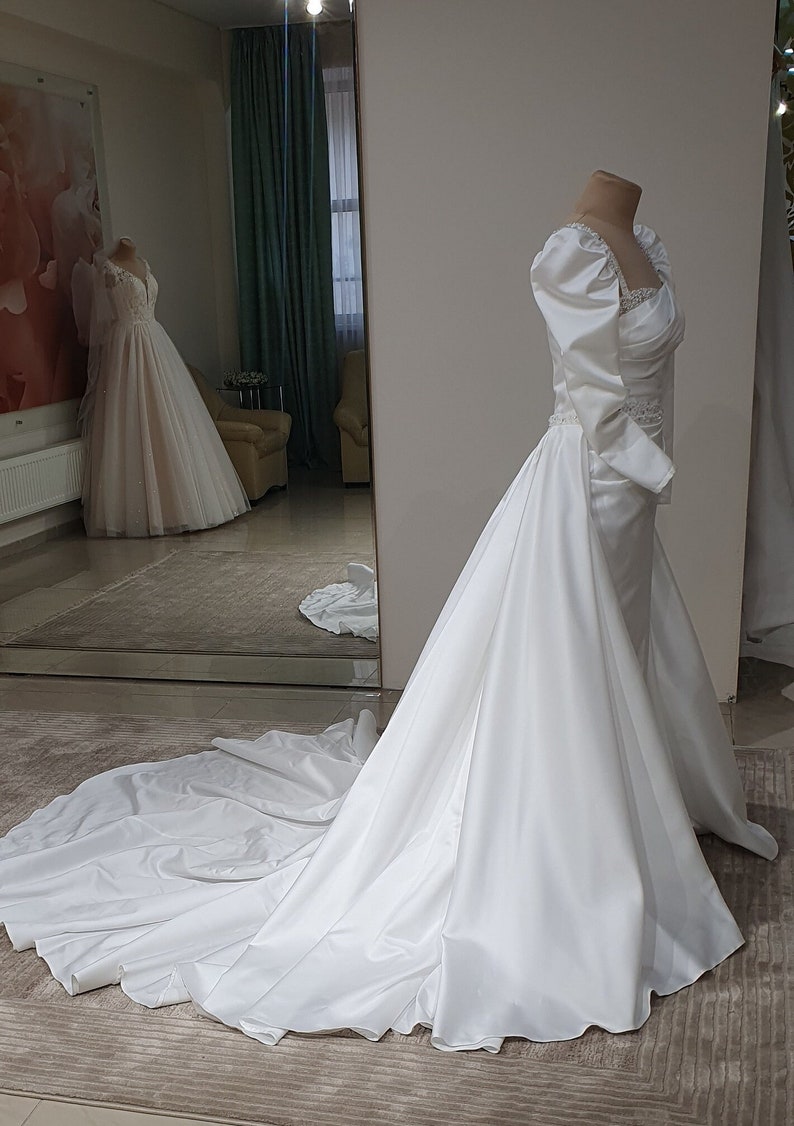 Puede incluir: Un vestido de novia de sat&eacute;n blanco con un corpi&ntilde;o ajustado y una falda fluida. El vestido tiene un escote de coraz&oacute;n y mangas largas con hombros abullonados. La falda tiene una cola y una sobrefalda desmontable.