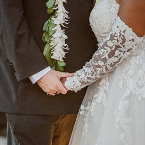 May include: A couple holding hands during a wedding ceremony. The groom is wearing a dark suit with a white flower lei. The bride is wearing a white lace wedding dress with a long veil.