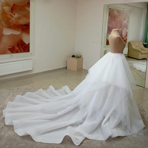 Puede incluir: Una falda de novia de tul blanco con una larga cola. La falda está en capas y tiene un dobladillo con volantes.