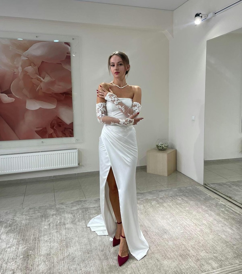 May include: A white satin wedding dress with a high slit and lace sleeves. The dress has a strapless neckline and a fitted bodice. The model is wearing red heels.