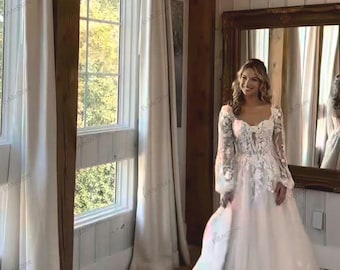 May include: A white wedding dress with long sleeves and floral lace detailing. The dress has a sweetheart neckline and a flowing skirt. The bride is standing in a room with a large window and a chandelier.