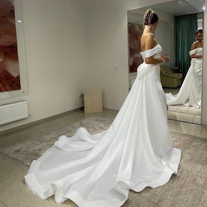 Puede incluir: Un vestido de novia blanco con una larga cola y un corpiño ajustado. El vestido tiene un escote de corazón y tirantes de hombros descubiertos. La novia está de pie en una habitación con un espejo y una alfombra.