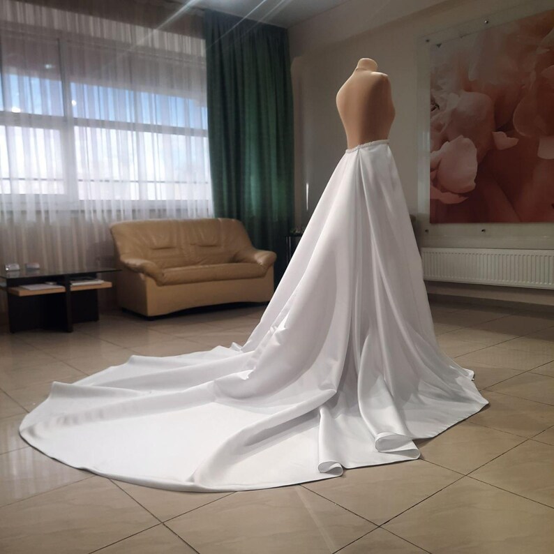 Puede incluir: Un vestido de novia blanco de sat&eacute;n con una larga cola. El vestido est&aacute; en un maniqu&iacute; y se exhibe en una habitaci&oacute;n con un sof&aacute; y una ventana.