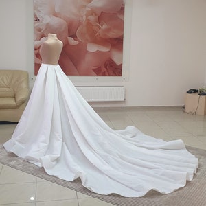 Puede incluir: Un vestido de novia blanco con una larga cola. El vestido tiene un corpiño ajustado y una falda completa. La falda está hecha de capas de tela que crean un efecto fluido.